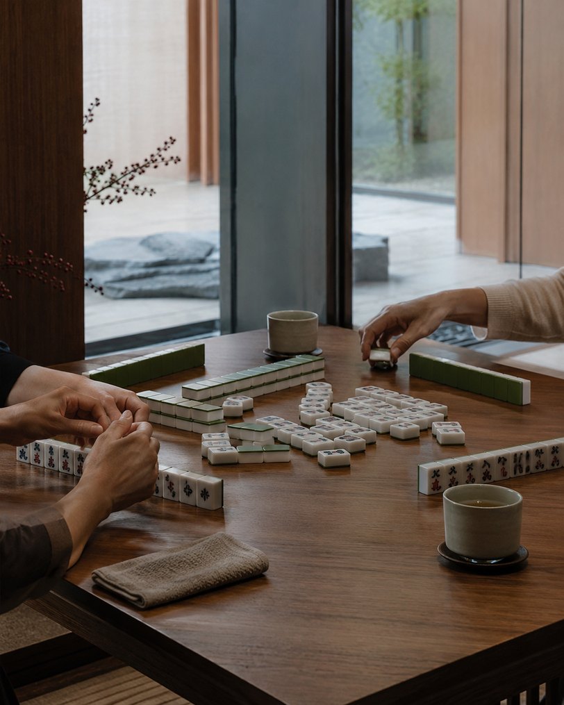 Hands arranging mahjong tiles on a wooden table with tea beside a courtyard window