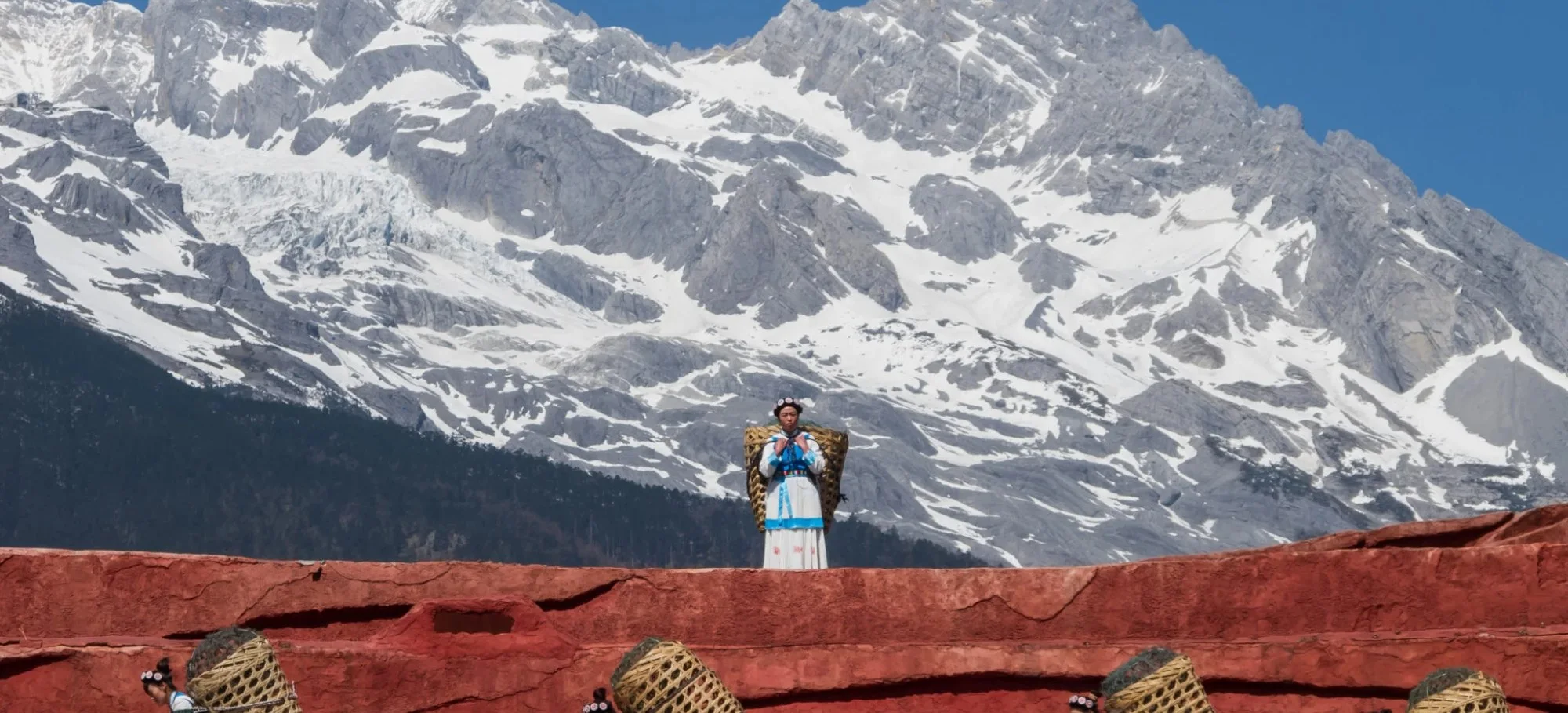 A woman in traditional dress standing on a red stone wall beneath a snow-capped mountain range, with a line of women carrying woven baskets below